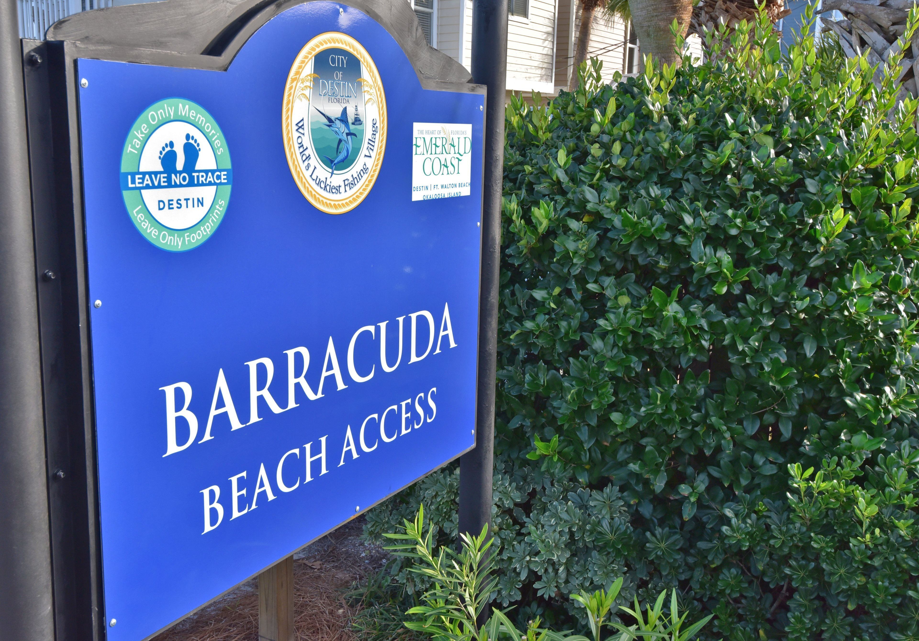 Un letrero azul con la inscripción 'BARRACUDA BEACH ACCESS' y logotipos de 'Take Only Memories' y 'World's Luckiest Fishing Village'.
