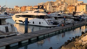 A marina with several yachts docked, a wooden pier, and buildings in the background.