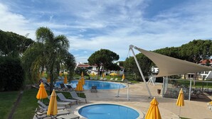 A pool area with sun loungers and umbrellas, surrounded by greenery and a clear sky.