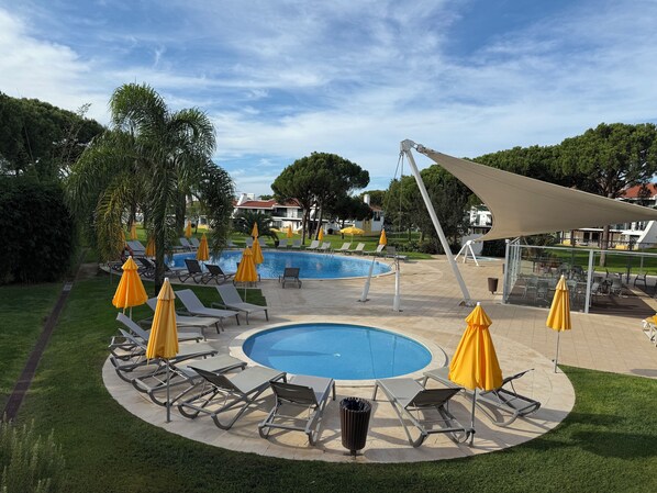 A pool area with sun loungers and umbrellas, surrounded by greenery and a clear sky.