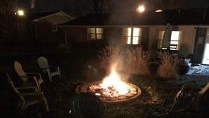 Een achtertuin met een vuurkorf, terrasstoelen en een huis met ramen en een deur.
