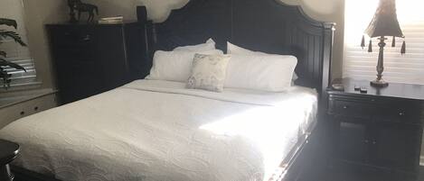 Una cama bien hecha con un cabecero oscuro, una mesita de noche con lámpara y un tocador con espejo.