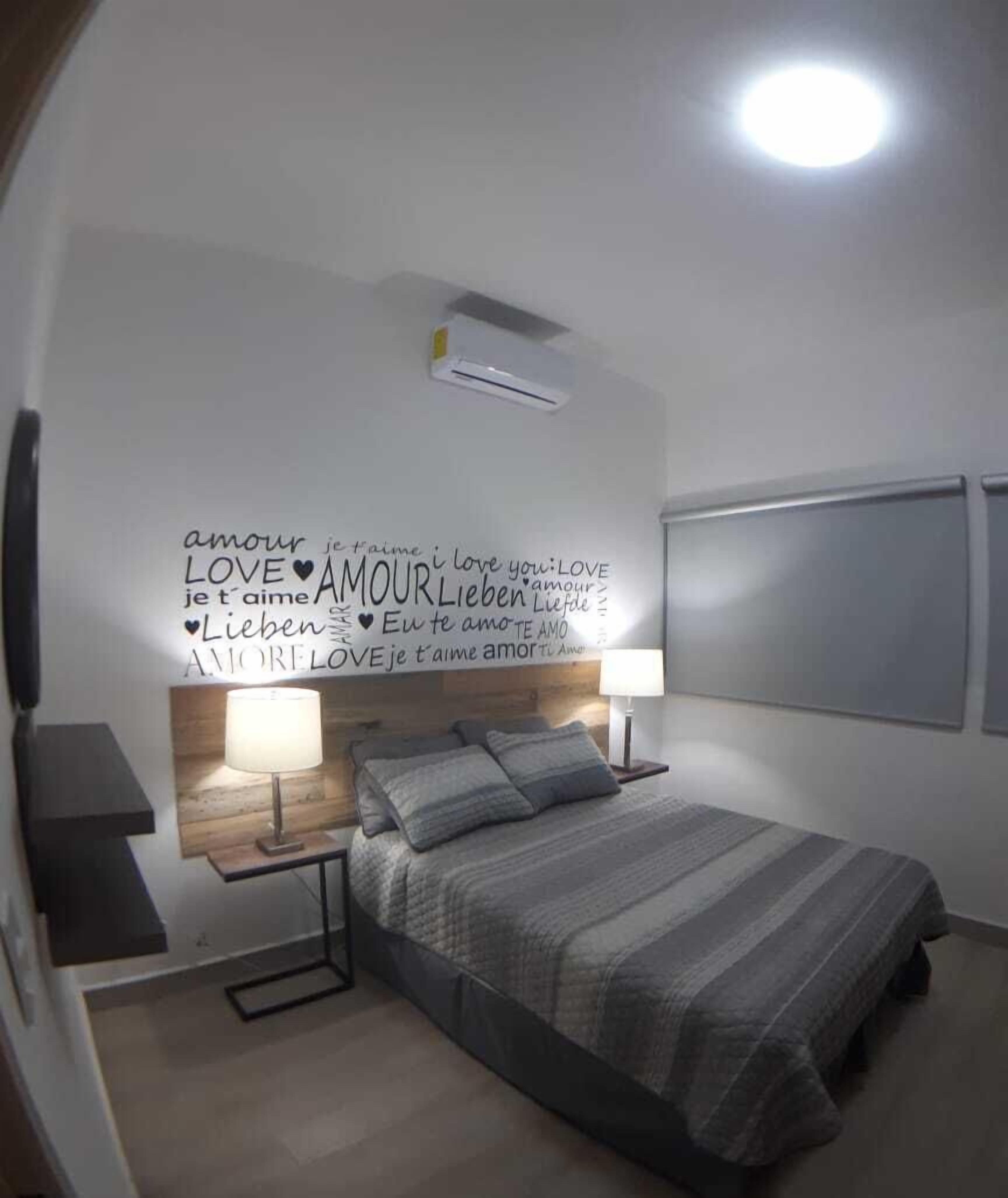 Un dormitorio moderno con cama, mesitas de noche, aire acondicionado de pared y un decorado con frases de amor en varios idiomas.