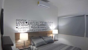 Un dormitorio moderno con cama, mesitas de noche, aire acondicionado de pared y un decorado con frases de amor en varios idiomas.
