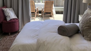 A bedroom with a large bed, a small red chair, a wooden table with chairs, and a view of a wooden fence and greenery outside. - Perfectly located, Modern, custom built casita w/AC & rooftop igloo sunset deck (San Diego)