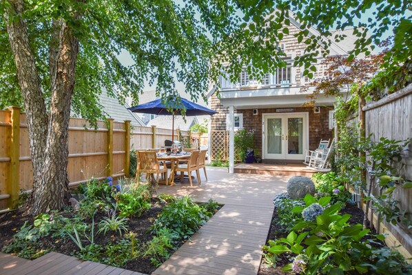 A backyard with a wooden deck, a dining table with chairs, a barbecue grill, and a covered patio area.