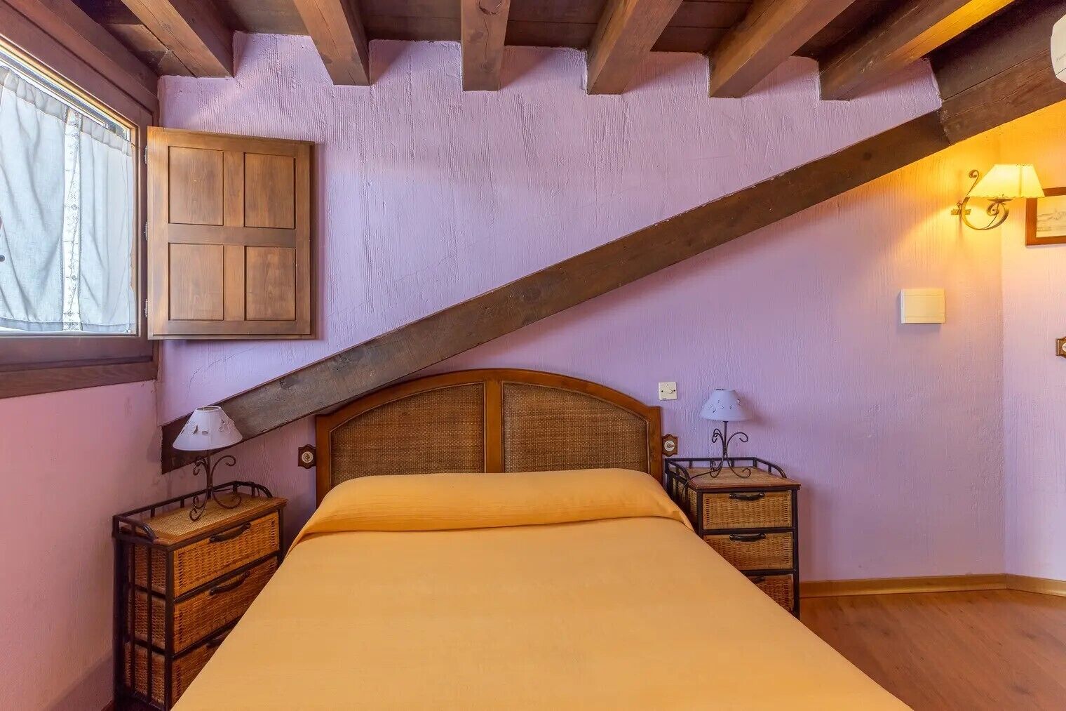 Een slaapkamer met een schuine kap, een houten bed, twee nachtkastjes met lampen, en een raam met gordijnen.