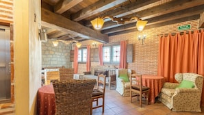 A rustic interior with exposed wooden beams, stone walls, and a brick fireplace. There are wicker chairs, a table, and a sign indicating an emergency exit. - Alval House for 23 people (Villacastín)