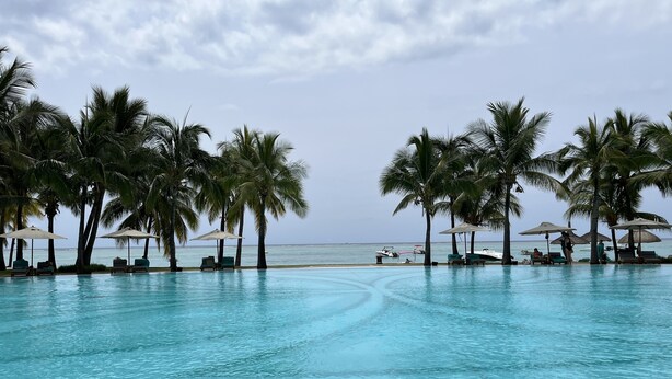 Zona junto a la piscina con sillones de descanso y sombrillas, palmeras y vista al océano.