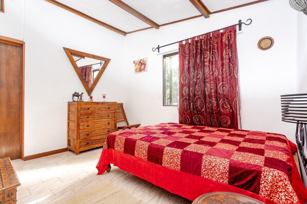Een slaapkamer met een houten dressoir, een rode geborduurde sprei, een spiegel en een raam met gordijnen.