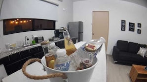 Keittiössä on valkoinen työtaso, pesuallas ja jääkaappi. Siellä on myös valkoinen ämpäri, jossa on erilaisia esineitä, kuten pulloja ja olutpullo. Olohuoneessa näkyy musta sohva ja puinen pöytä.