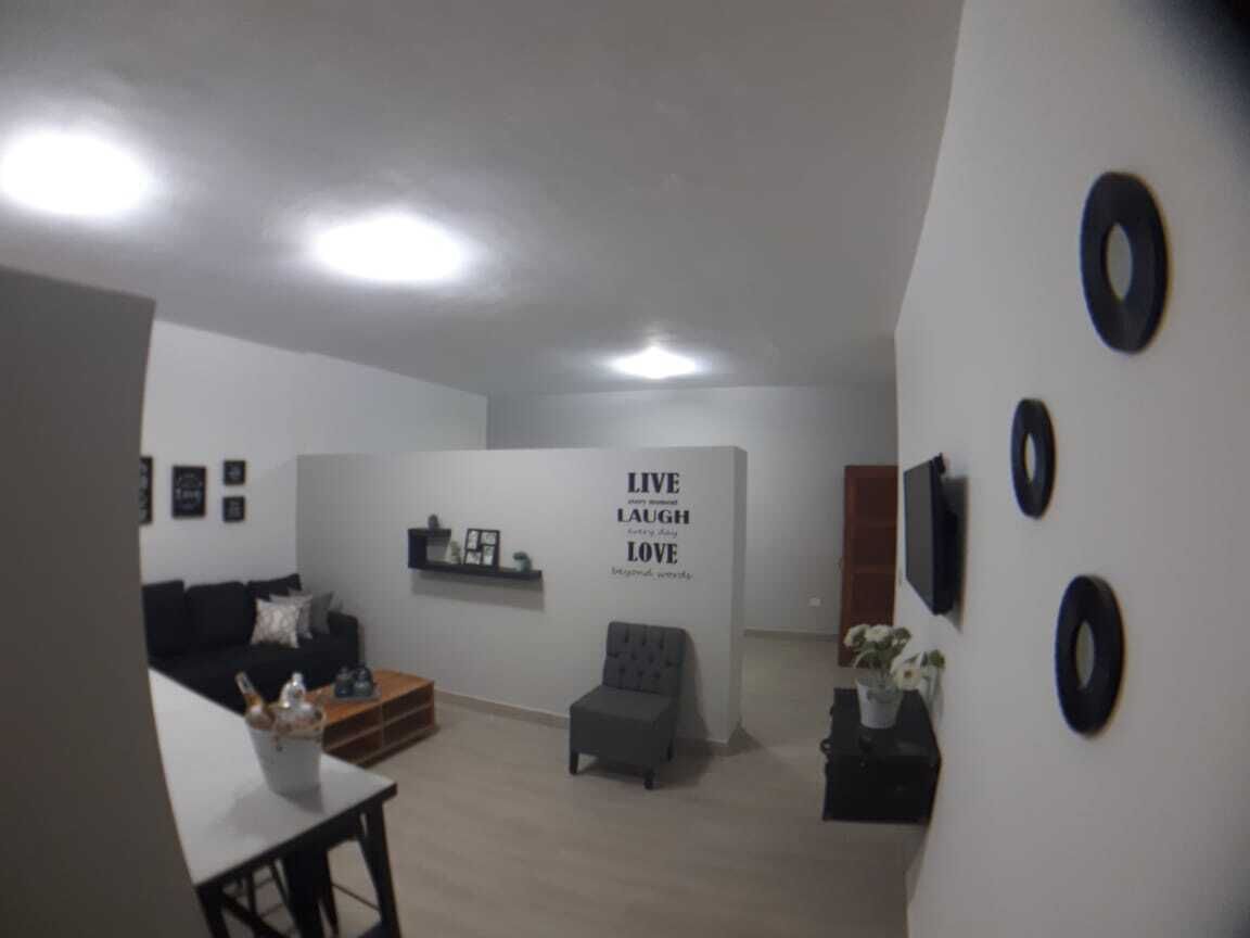 En moderne stue med en sort sofa, en grå skammel og et sofabord. Væggen har et motiverende citat, og en lille hylde viser dekorative genstande.