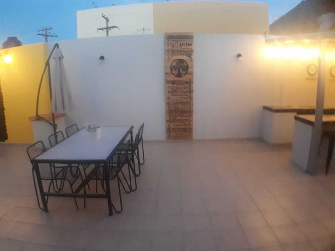 Um pátio externo com uma mesa retangular e cadeiras, uma parede de madeira com um letreiro vertical e uma pequena estrutura com iluminação.