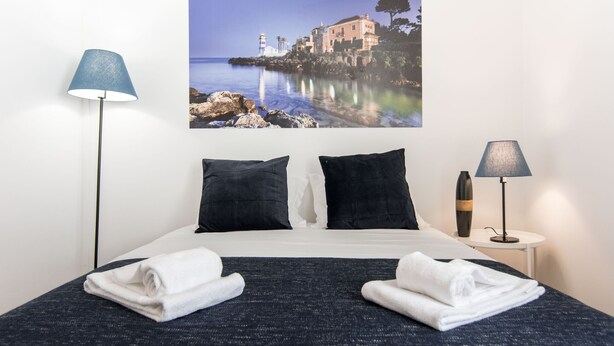 A neatly made bed with two pillows, two folded towels, and a painting of a coastal scene above it. - Casa Sanches (Cascais)