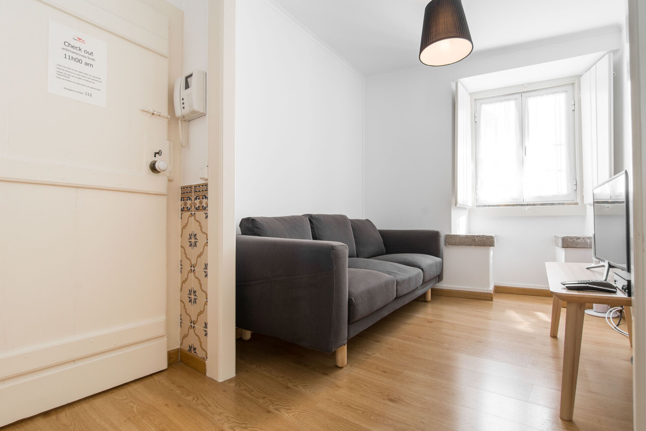 Ein modernes Wohnzimmer mit einem grauen Sofa, Holzboden, einem Schreibtisch mit Computer und einem Fenster mit weißen Fensterläden.