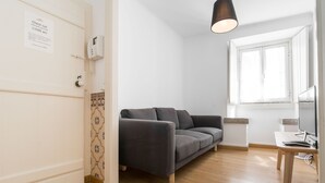 A modern living room with a grey sofa, wooden flooring, a desk with a computer, and a window with white shutters. - Casa Sanches (Cascais)