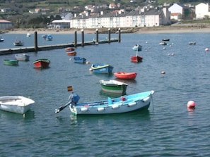 A harbor with various boats anchored near the shore, surrounded by buildings and a sandy beach.