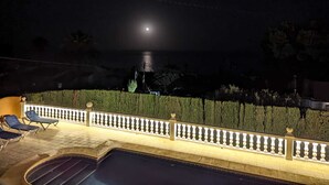 A nighttime pool area with a moonlit view over the water, a tiled deck, and a white balustrade. - Villa with panoramic views of the sea and the Rock of Ifach, with private pool. (Calpe)