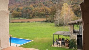 A view through an arched window of a pool, a patio with outdoor furniture, and a hillside with autumn foliage.