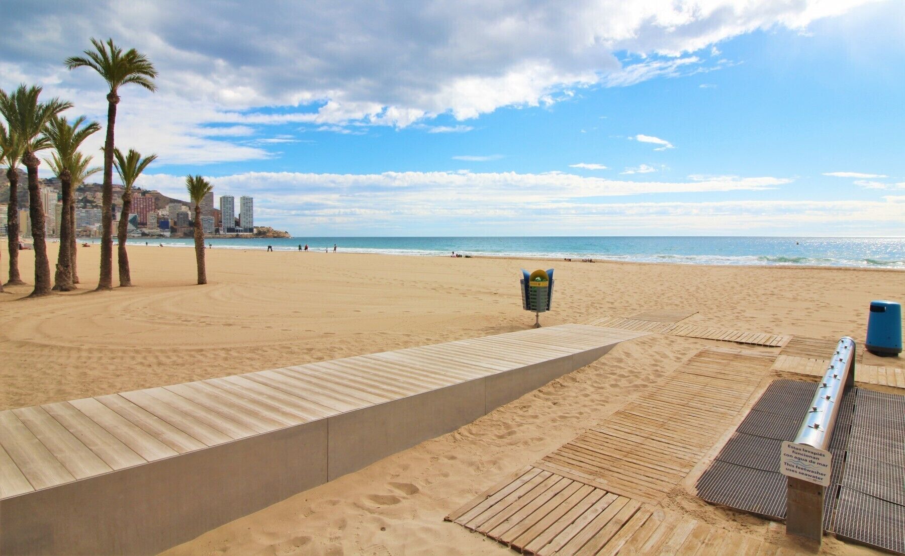 A beach with a wooden walkway, palm trees, and a cityscape in the background.