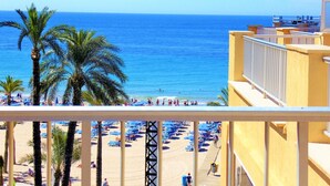 A beach with palm trees, a clear blue sky, and a building with a balcony overlooking the beach. - Atico Ancla Penthouse (Benidorm)