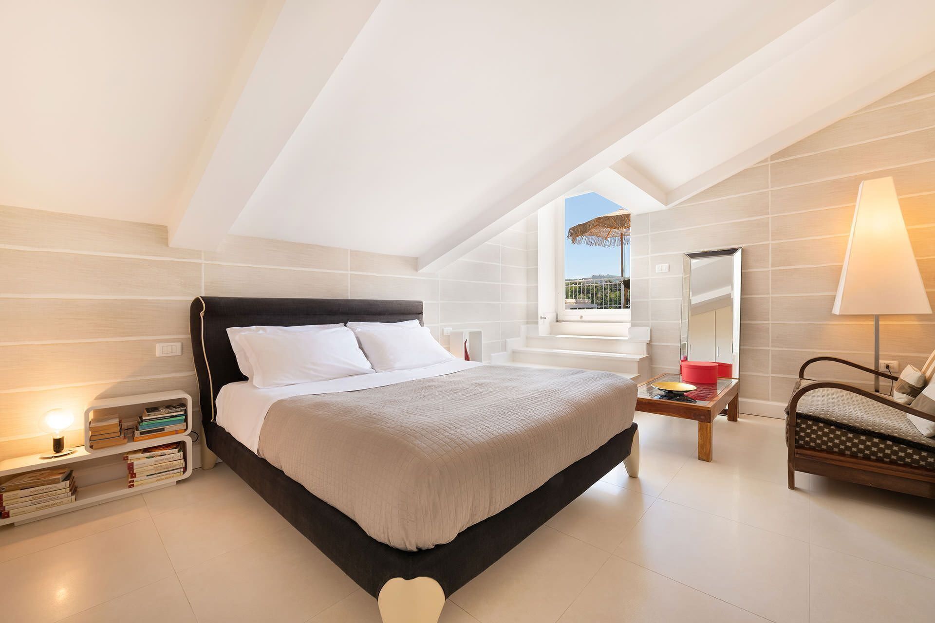 A modern bedroom with a large bed, a bedside table with books, a chair, a small table with a lamp, and a window with a view of the outside.