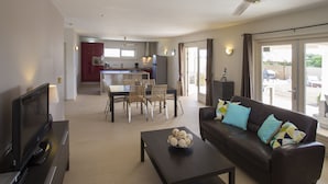 A modern living room with a flat-screen TV, a dark brown sofa with colorful cushions, a coffee table with decorative items, and a dining area with wicker chairs and a table.