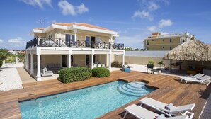 A two-story house with a swimming pool, lounge chairs, and a thatched-roof structure.