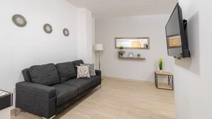 En moderne stue med en grå sofa, et fladskærms-tv monteret på væggen, et lille sofabord med en plante og et spejl over en hylde med pynt.