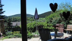 Une terrasse extérieure offrant une vue sur un paysage verdoyant, avec des collines et des arbres. On y trouve deux chaises et une table sur laquelle est posée une plante en pot.