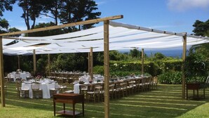 Un espace extérieur aménagé pour les événements, avec des tables disposées sous une tonnelle blanche, entouré de verdure et d’arbres.