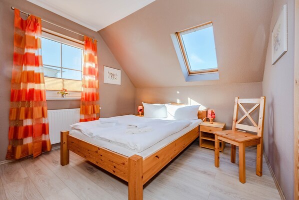 Un dormitorio con una cama de madera, una mesita de noche con lámpara, una pequeña silla de madera, una ventana con cortinas y un tragaluz.