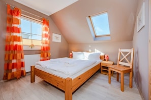 Une chambre avec un lit en bois, une table de chevet avec une lampe, une petite chaise en bois, une fenêtre avec des rideaux et un puits de lumière.