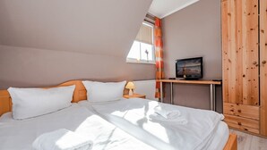 Un lit bien fait, avec du linge de lit blanc et une serviette pliée, une table de chevet avec une lampe et un téléviseur posé sur un meuble en bois.
