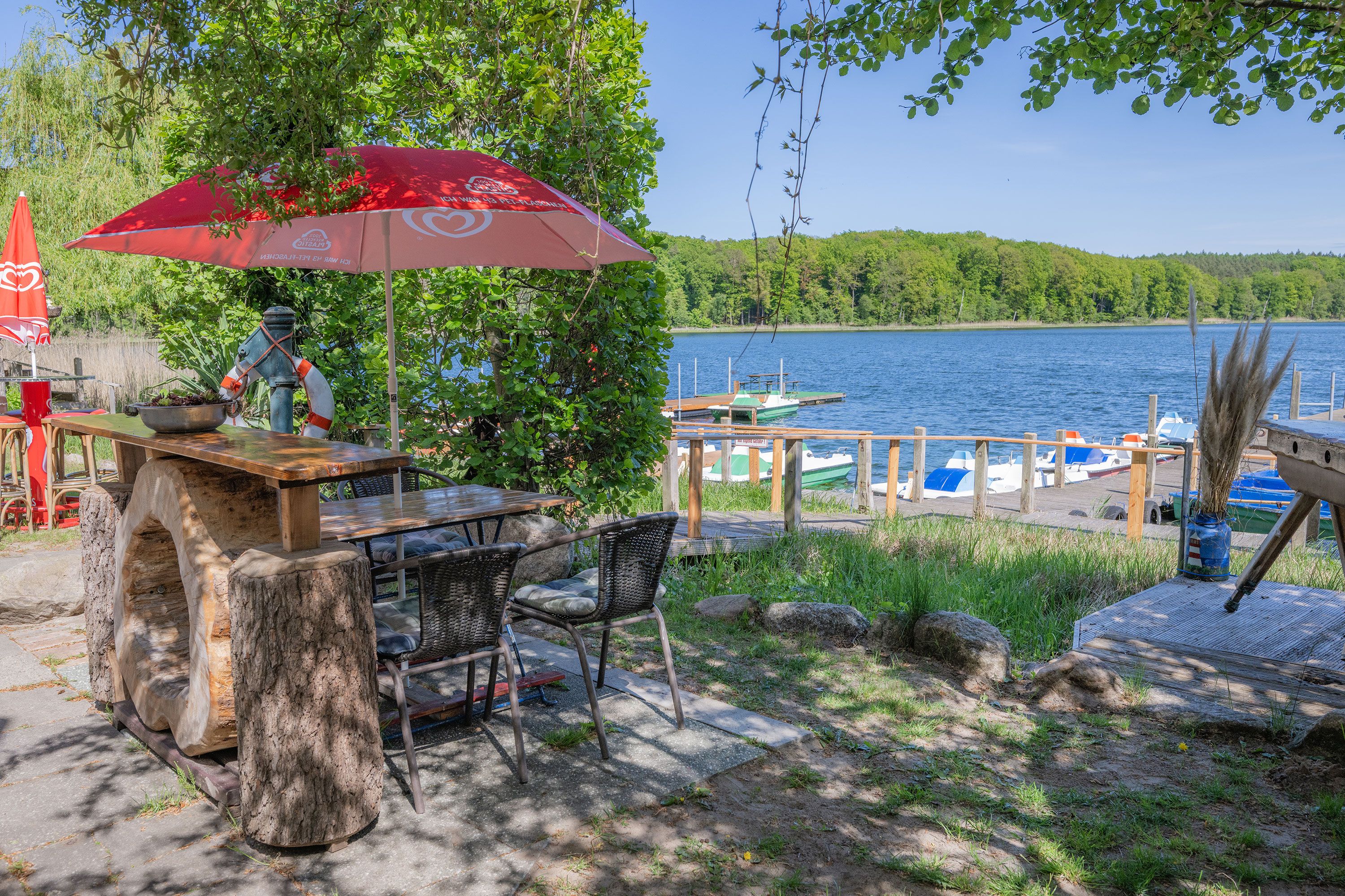 An outdoor dining area with a table and chairs, a red umbrella, and a view of a lake with boats.