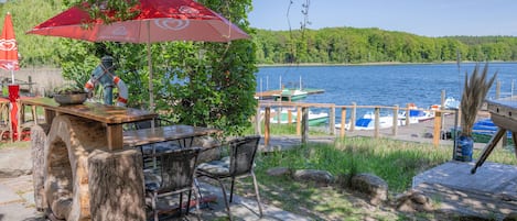 Un espace extérieur aménagé pour les repas, avec une table et des chaises, un parasol rouge et une vue sur un lac où naviguent des bateaux.