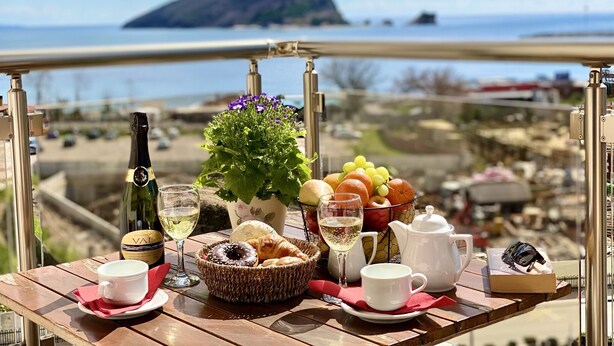 Une table dressée avec une bouteille de vin, un verre, un panier de croissants, un saladier de fruits, une théière et une tasse de café, offrant une vue panoramique sur une montagne et la mer.