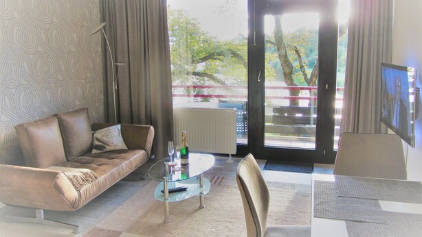 Ein Wohnzimmer mit einer Couch, einem Couchtisch mit Flasche und Gläsern, einem Fernseher auf einem Schrank und Blick auf Bäume und einen Balkon.