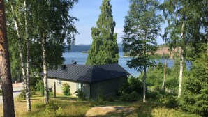 A small cabin with a metal roof, surrounded by trees and a lake view. - Idyllic vacation home right by the sea<br> (Docksta)