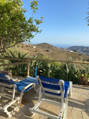 Eine Terrasse mit zwei blauen und weißen Liegestühlen, einer Holz-Pergola und Blick auf eine hügelige Landschaft unter blauem Himmel.