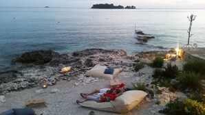A rocky beach with two people lying on cushions, a small wooden bench, and a distant island. - Apartment Lella near the sea and city center 500 meters (Rovinj)