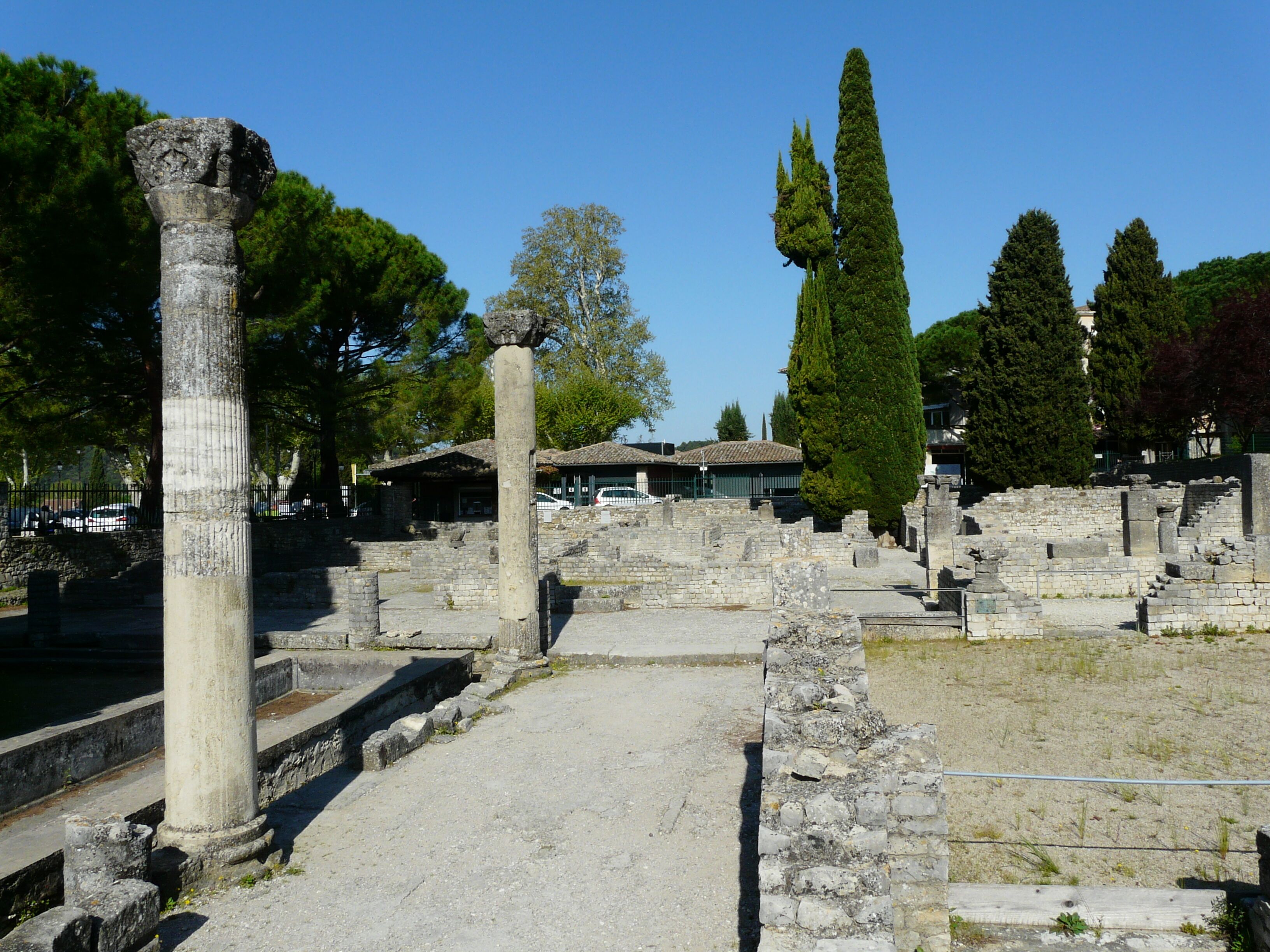 Ancient ruins with stone columns and pathways, surrounded by trees and a building in the background.
