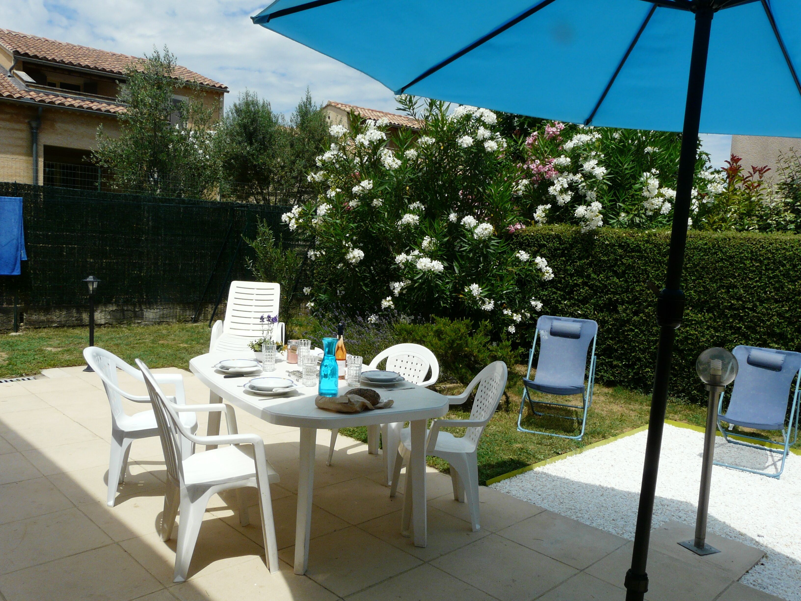 An outdoor patio with a table set for four, surrounded by white plastic chairs, under a blue umbrella.