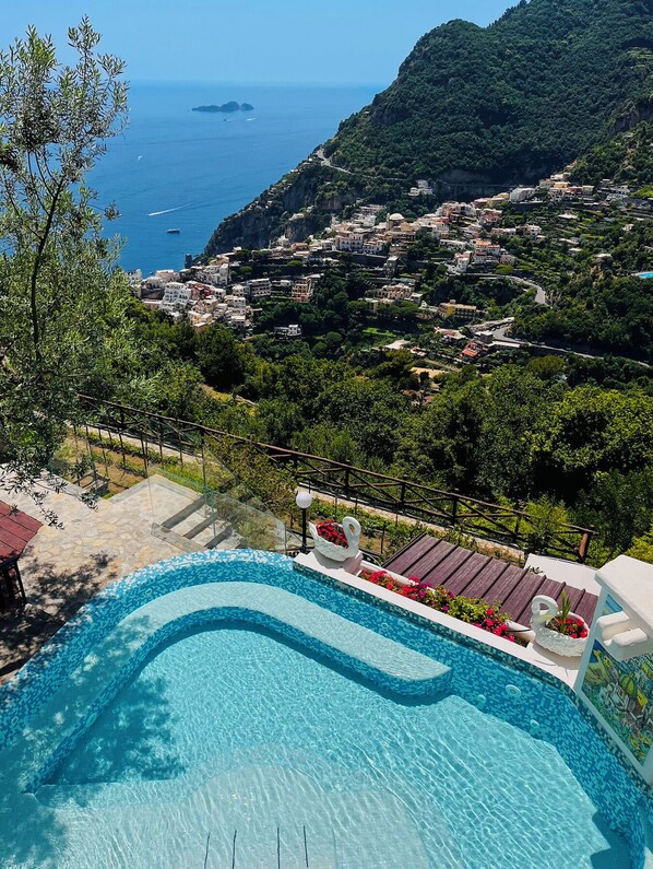 A swimming pool with clear blue water, surrounded by a stone deck and a white swan-shaped planter. In the background, there is a coastal village with buildings nestled among green hills and the sea.