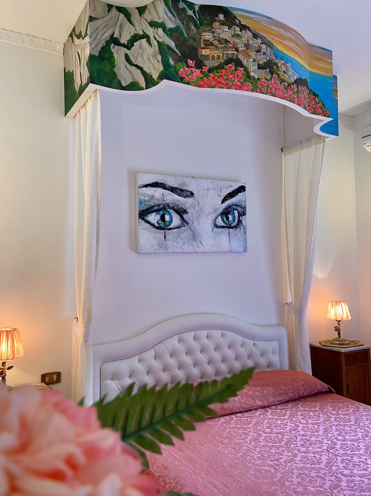 A bedroom with a white canopy bed, a painting of eyes on the wall, and a pink patterned bedspread.