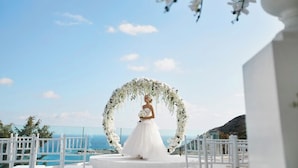 Hochzeitszeremonie mit Braut im weißen Kleid, einem Blumenschmuckbogen und Blick aufs Meer.
