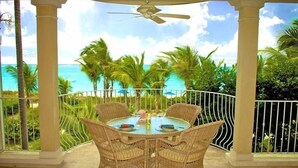 Un área de comedor al aire libre techada, con sillas de mimbre y una mesa, con vistas a una playa con palmeras y el océano.