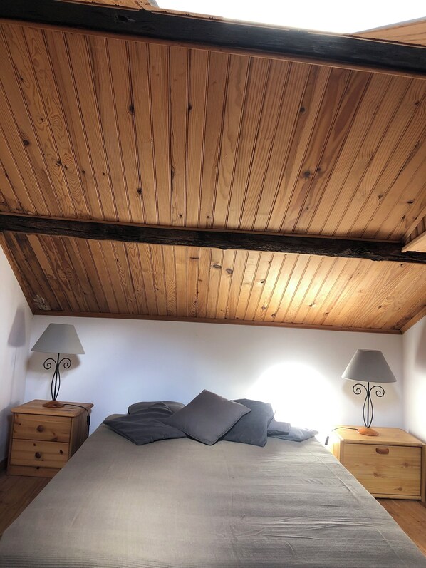 Ein Schlafzimmer mit Holzboden, einem Bett mit grauer Bettwäsche, zwei Nachttischen mit Lampen und einer kleinen Holzkammer.