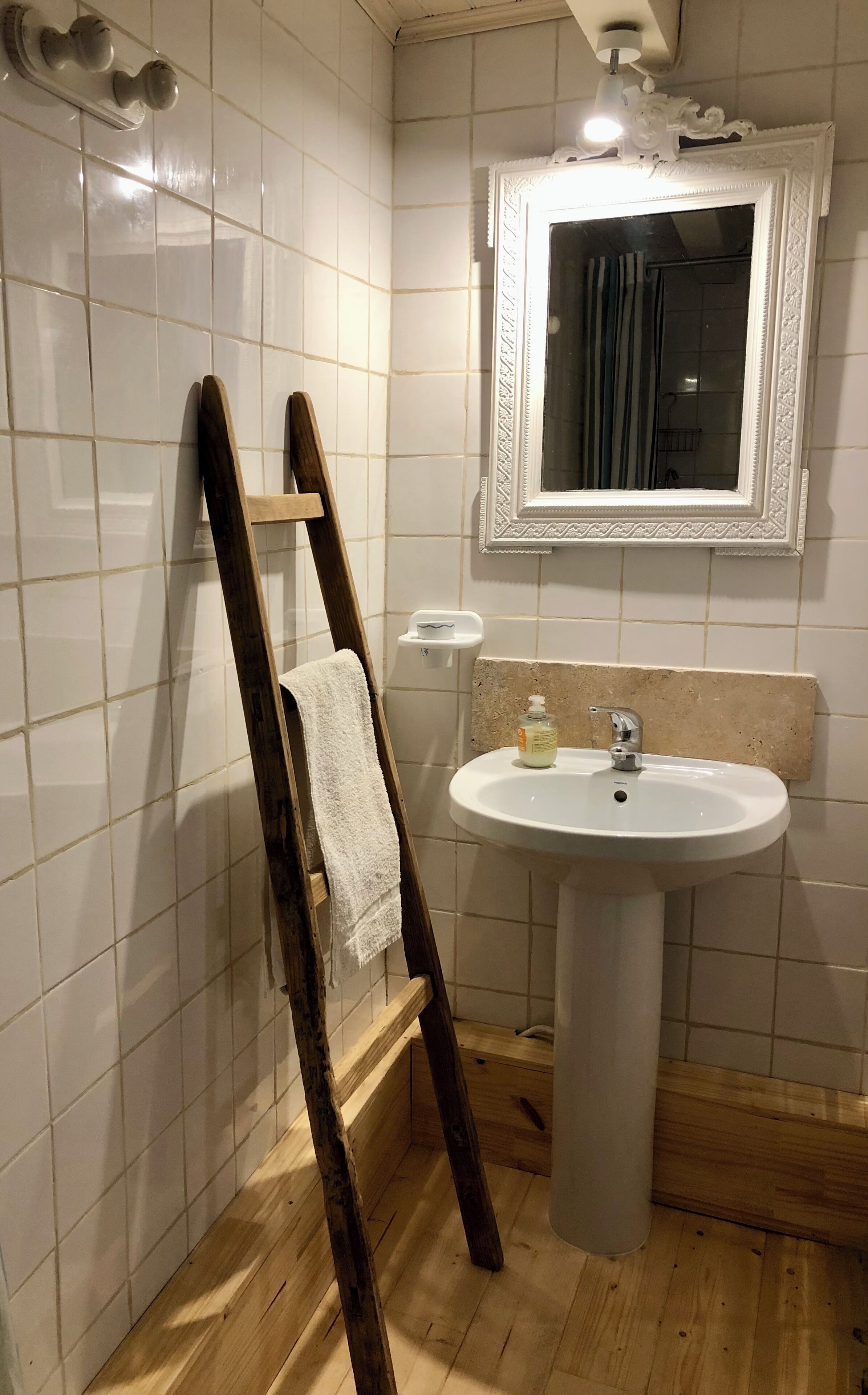 Et badeværelse med hvide flisevægge, en træstige lænet op ad væggen, et hvidt håndklæde hængende på stigen, en hvid vask med et spejl over, og en lysarmatur i loftet.