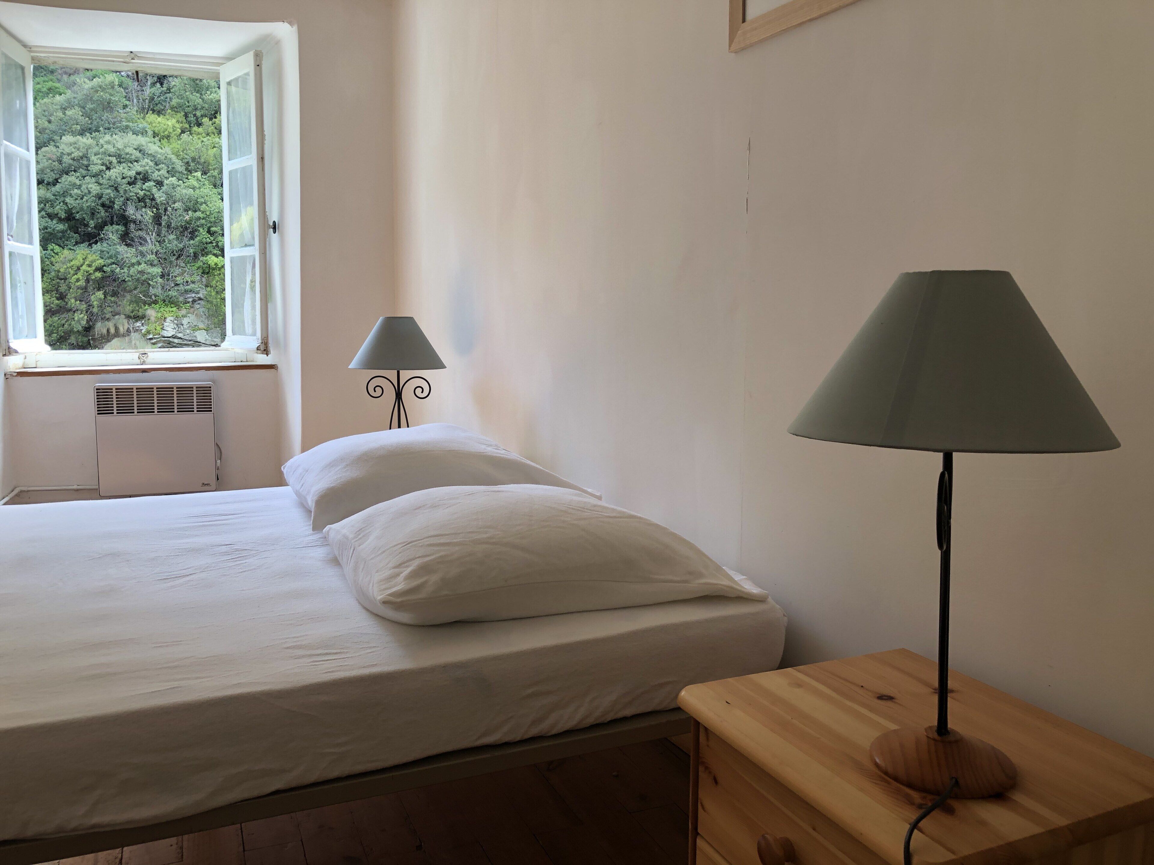 Een slaapkamer met een bed, een raam met uitzicht op bomen, een radiator en een lamp op een houten nachtkastje.